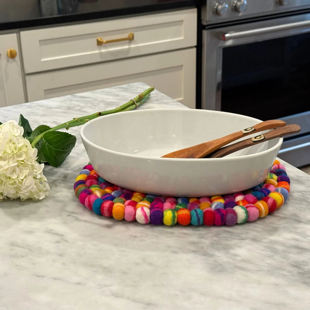 A white oval serving bowl with wooden serving utensils, placed on a colorful round felt placemat on a marble countertop in a kitchen setting.